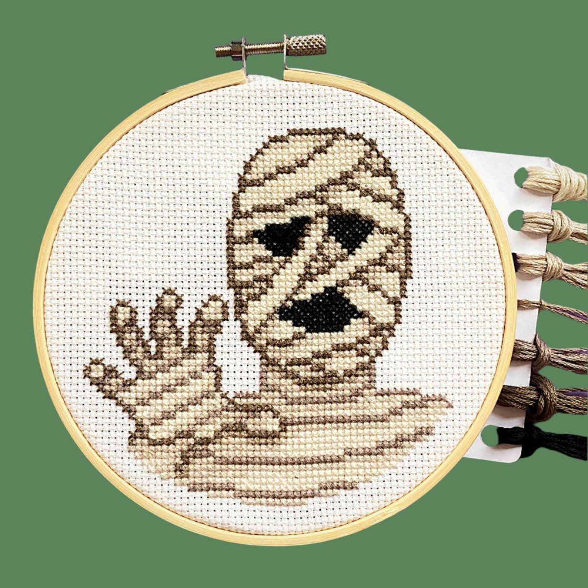 Cross stitch in embroidery hoop featuring bandaged mummy head with hand reaching out