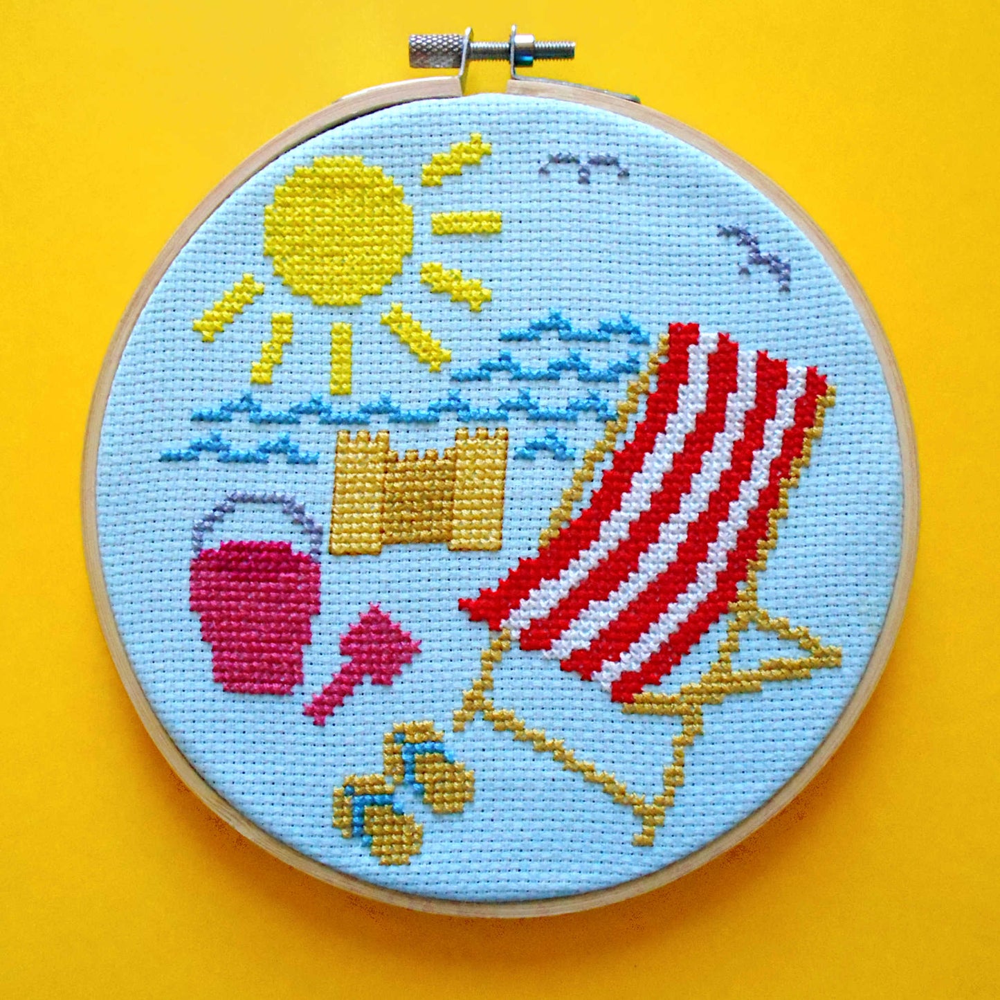 Cross stitch in embroidery hoop featuring beach scene - deckchair, flip flips, bucket and spade, sandcastle, sea, sun, seagulls on blue fabric