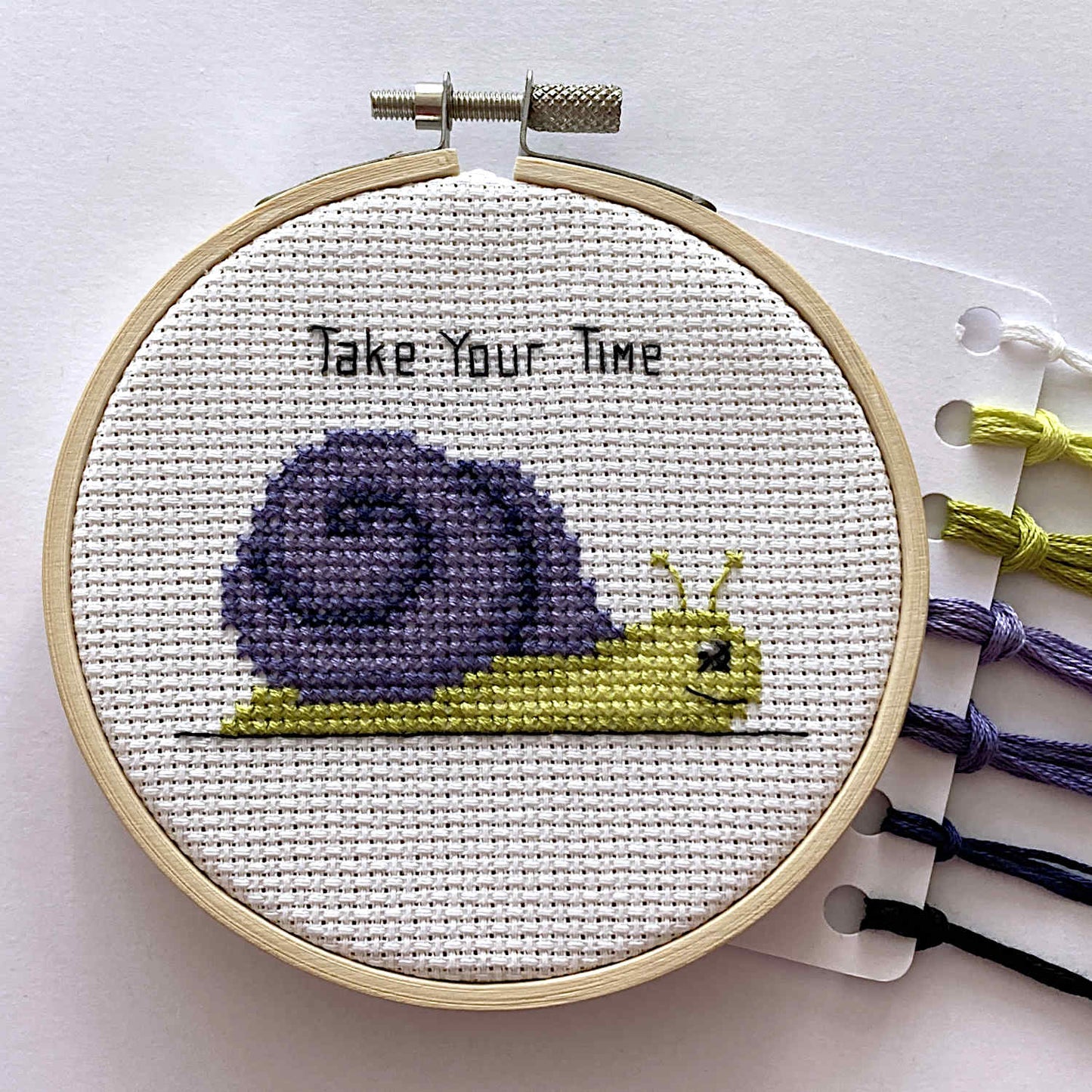 A completed cross-stitch piece in a wooden embroidery hoop, featuring a purple and yellow snail with the text 'Take Your Time' stitched above it. To the right, thread spools in purple, yellow, black, and white are visible on a white background.