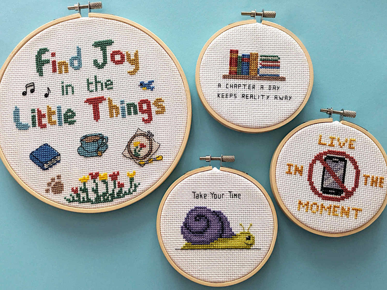 4 completed cross stitches in embroidery hoops with slogans 'Find Joy in the Little things', 'A Chapter a Day Keeps Reality Away', 'Take Your Time' and 'Live in the Moment'