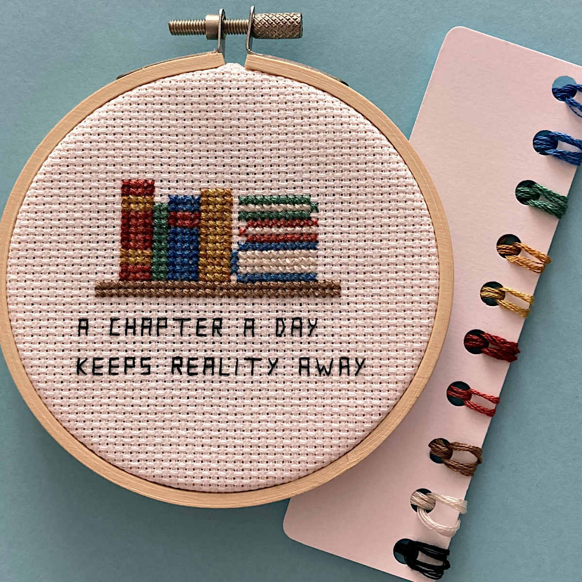 A completed cross-stitch embroidery in a wooden hoop showing colorful book spines and the text 'A CHAPTER A DAY KEEPS REALITY AWAY'. A thread card with multiple colored threads is placed beside it on a light blue background.