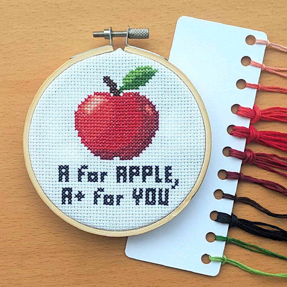 Cross-stitched apple design with text 'A for Apple, A+ for You' on a wooden surface with embroidery threads.