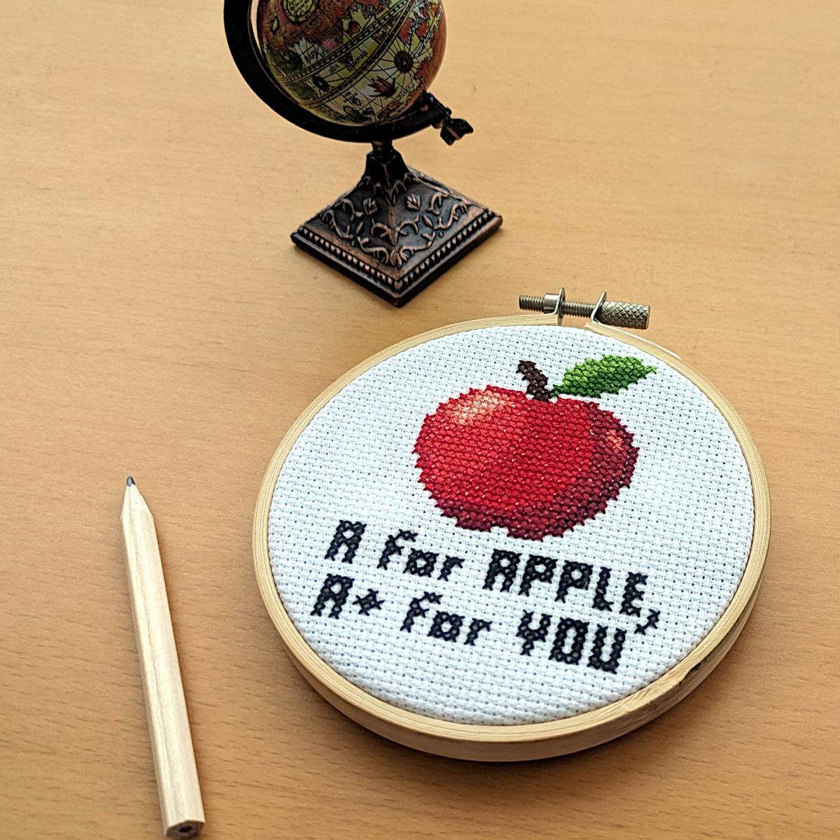 Cross-stitched apple design with text 'A for Apple, A+ for You' underneath in an embroidery hoop on a wooden surface.