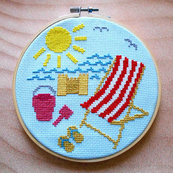 Cross stitch in embroidery hoop featuring beach scene - deckchair, flip flips, bucket and spade, sandcastle, sea, sun, seagulls on blue fabric