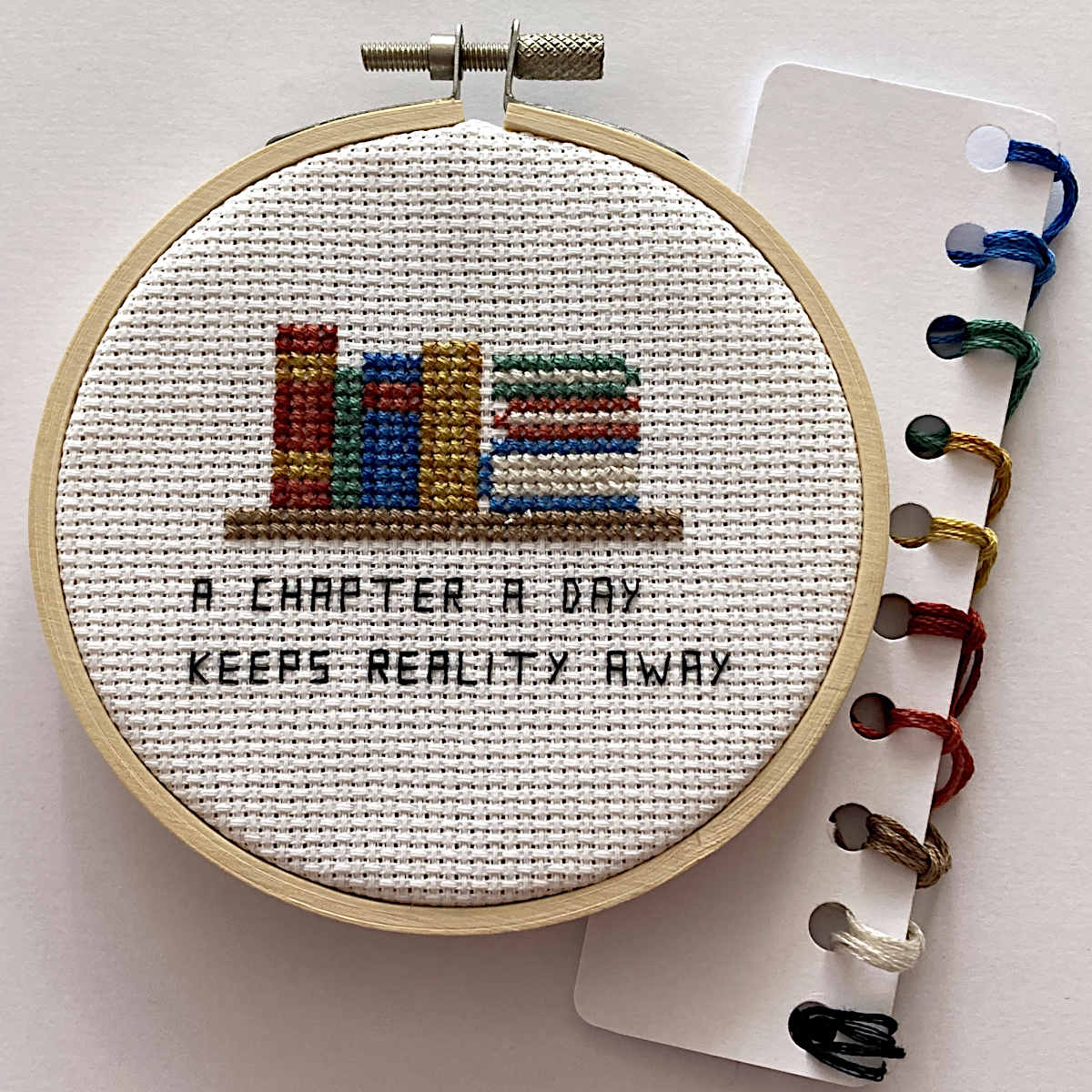A round embroidery hoop containing a cross-stitch design of colorful books and the text 'A CHAPTER A DAY KEEPS REALITY AWAY' on white fabric. A card with multiple thread colors attached is placed to the right on a light background.