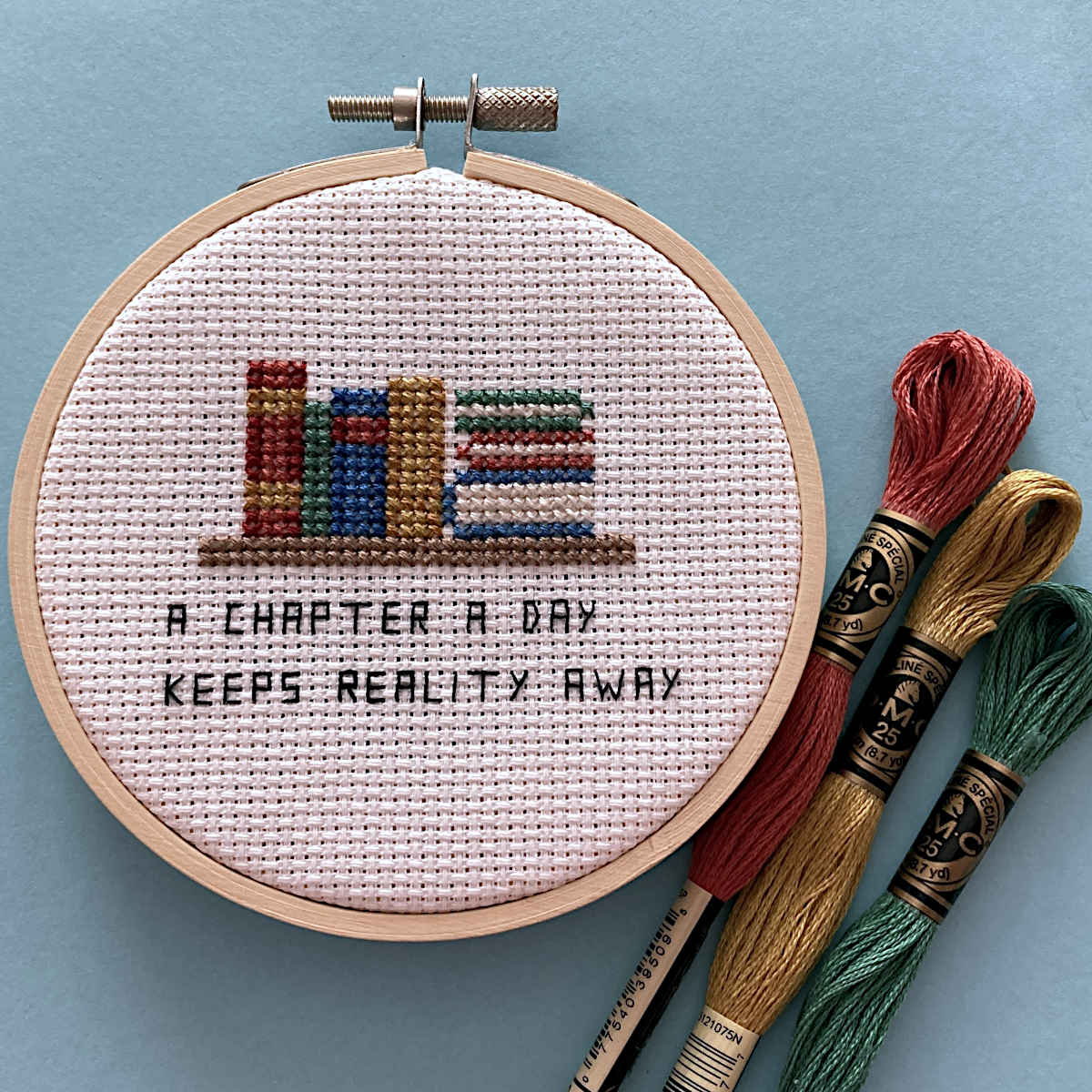 A cross-stitch embroidery hoop displaying a finished design of colorful books and the text 'A CHAPTER A DAY KEEPS REALITY AWAY'. Three skeins of embroidery floss in red, gold, and green are arranged next to the hoop on a light blue background.