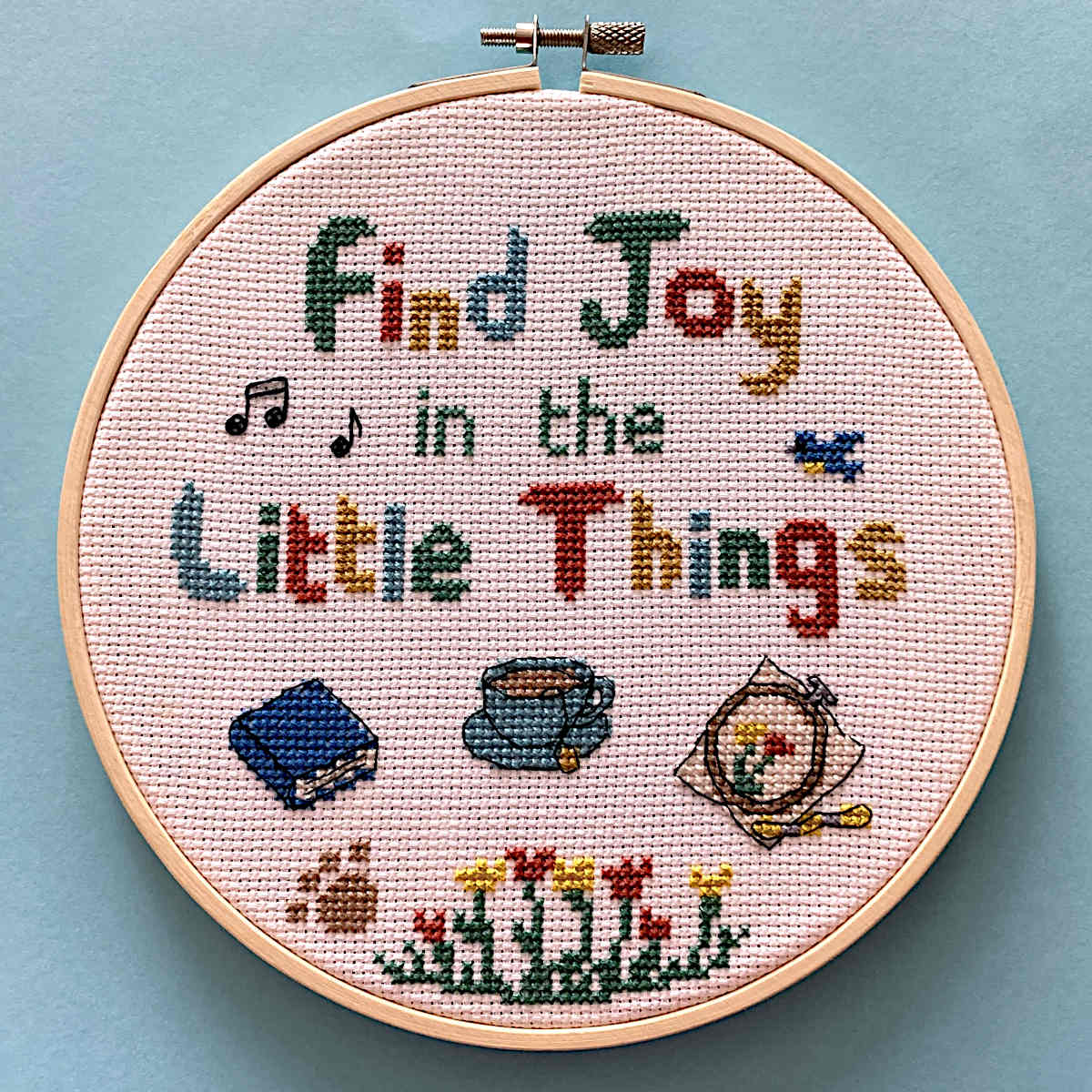 A circular cross-stitch embroidery hoop containing a completed cross-stitch design with the text 'Find Joy in the Little Things' and small cross stitched motifs including music notes, a bird, a book, a cup, embroidery hoop and flowers, set against a light blue background.