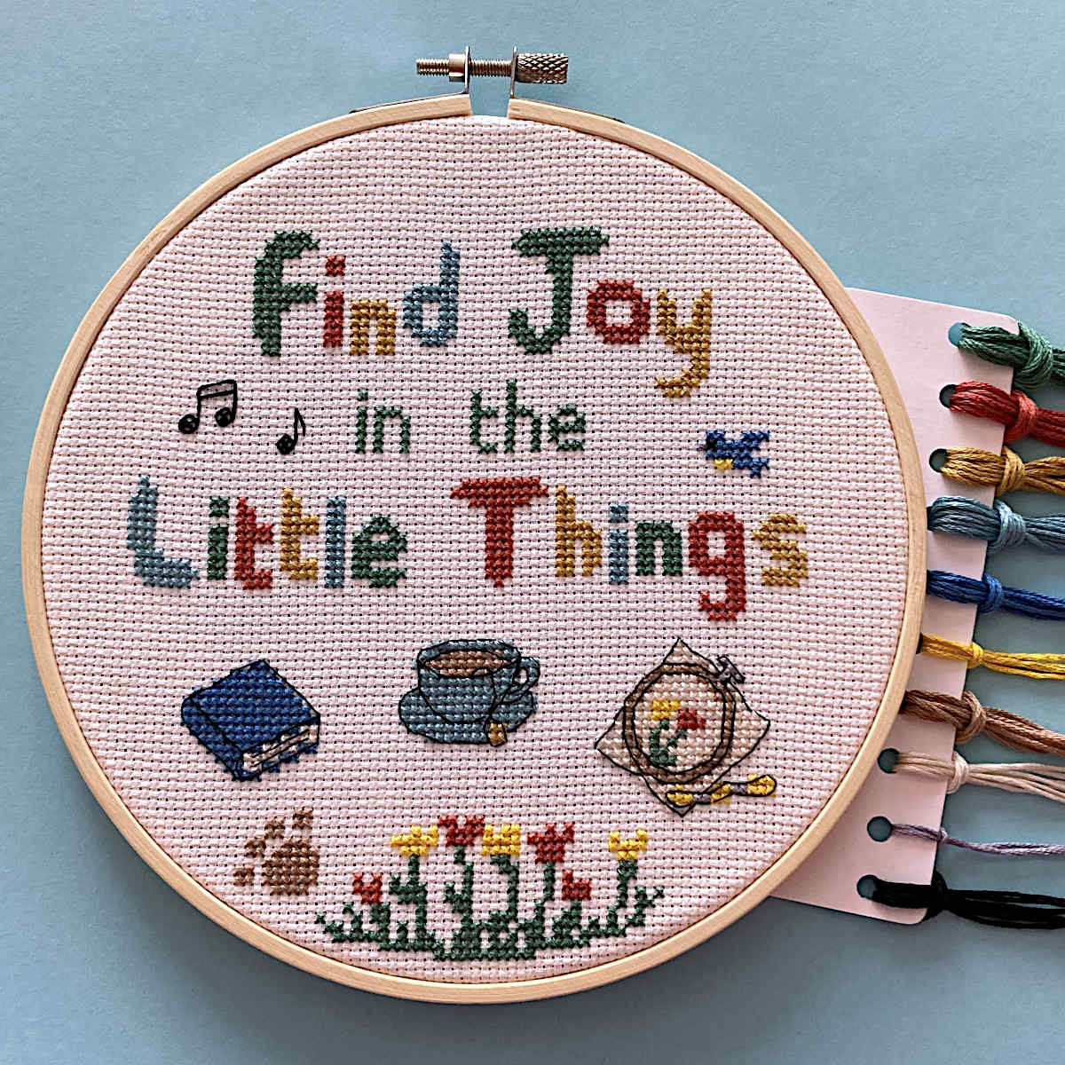 Cross stitch with 'Find Joy in the Little Things' text and small motifs of a book, a cup of tea, an embroidery and thread skein, flowers, paw print, musical notes, bird