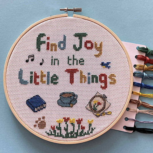 Cross stitch with 'Find Joy in the Little Things' text and small motifs of a book, a cup of tea, an embroidery and thread skein, flowers, paw print, musical notes, bird