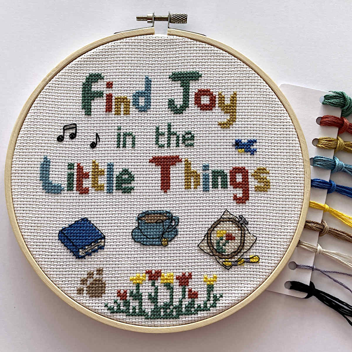 Cross stitch with 'Find Joy in the Little Things' text and small motifs of a book, a cup of tea, an embroidery and thread skein, flowers, paw print, musical notes, bird