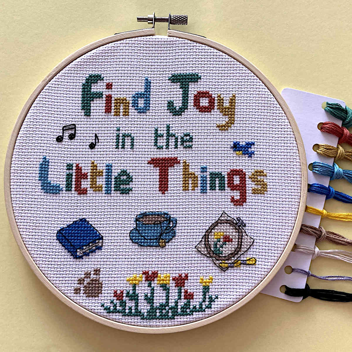 A completed cross-stitch piece in a wooden hoop on a pale yellow background, with a card of thread spools partially visible on the right. The stitched design reads “Find Joy in the Little Things” in multicolored letters, with small embroidered motifs including music notes, a blue book, a cup, a bird, flowers, and a paw print.
