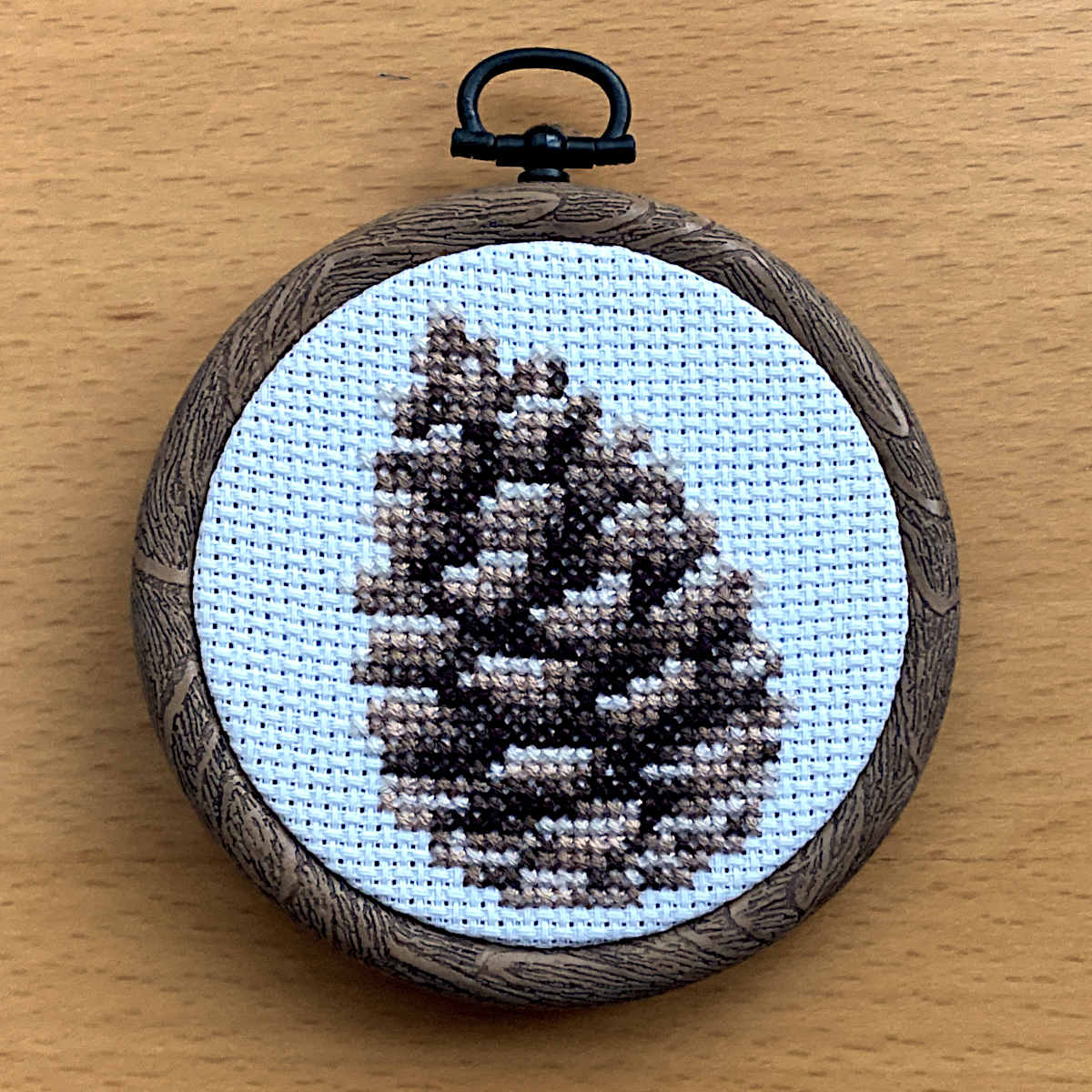 A completed pinecone cross stitch on white fabric, displayed in a round embroidery hoop 