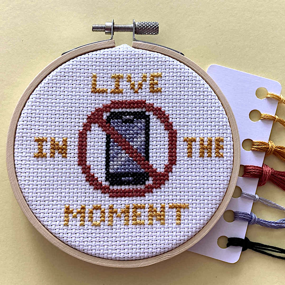 A completed cross-stitch piece in a wooden hoop on a pale yellow background, with a card of embroidery floss threads partially visible on the right. The design reads “LIVE IN THE MOMENT” around a crossed-out phone icon.
