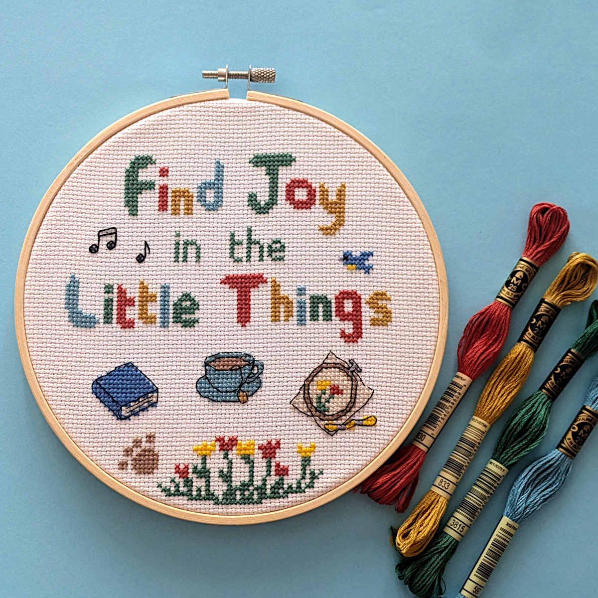 A round embroidery hoop with a completed cross-stitch design that reads 'Find Joy in the Little Things' in multicolored letters, surrounded by small stitched motifs including music notes, a book, a teacup, flowers, and an embroidery hoop. Several DMC embroidery floss skeins in red, gold, green, blue, and brown are arranged beside it on a light blue background.