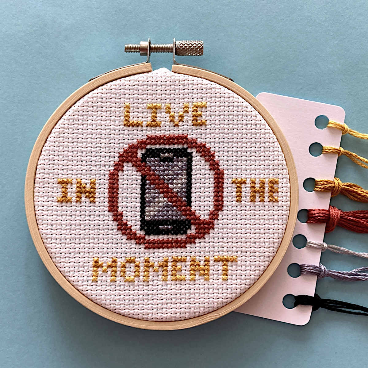 A circular embroidery hoop containing a cross-stitch design that reads 'LIVE IN THE MOMENT' around a crossed-out phone icon. A white thread card with DMC embroidery thread is partially visible on the right against a light blue background.
