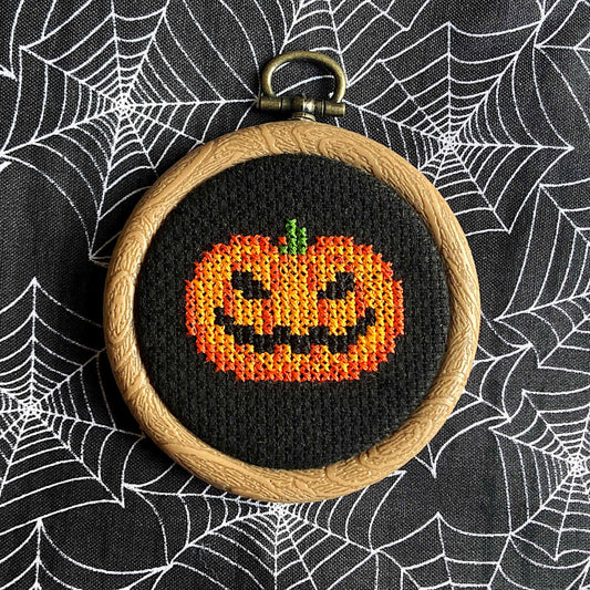Mini orange jack o'lantern/pumpkin cross stitch on black fabric, in embroidery hoop