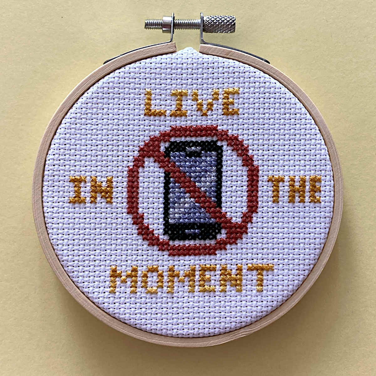 A circular embroidery hoop containing a cross-stitch design that reads 'LIVE IN THE MOMENT' with a red prohibition symbol over a smartphone. The hoop is placed against a solid pale yellow background.