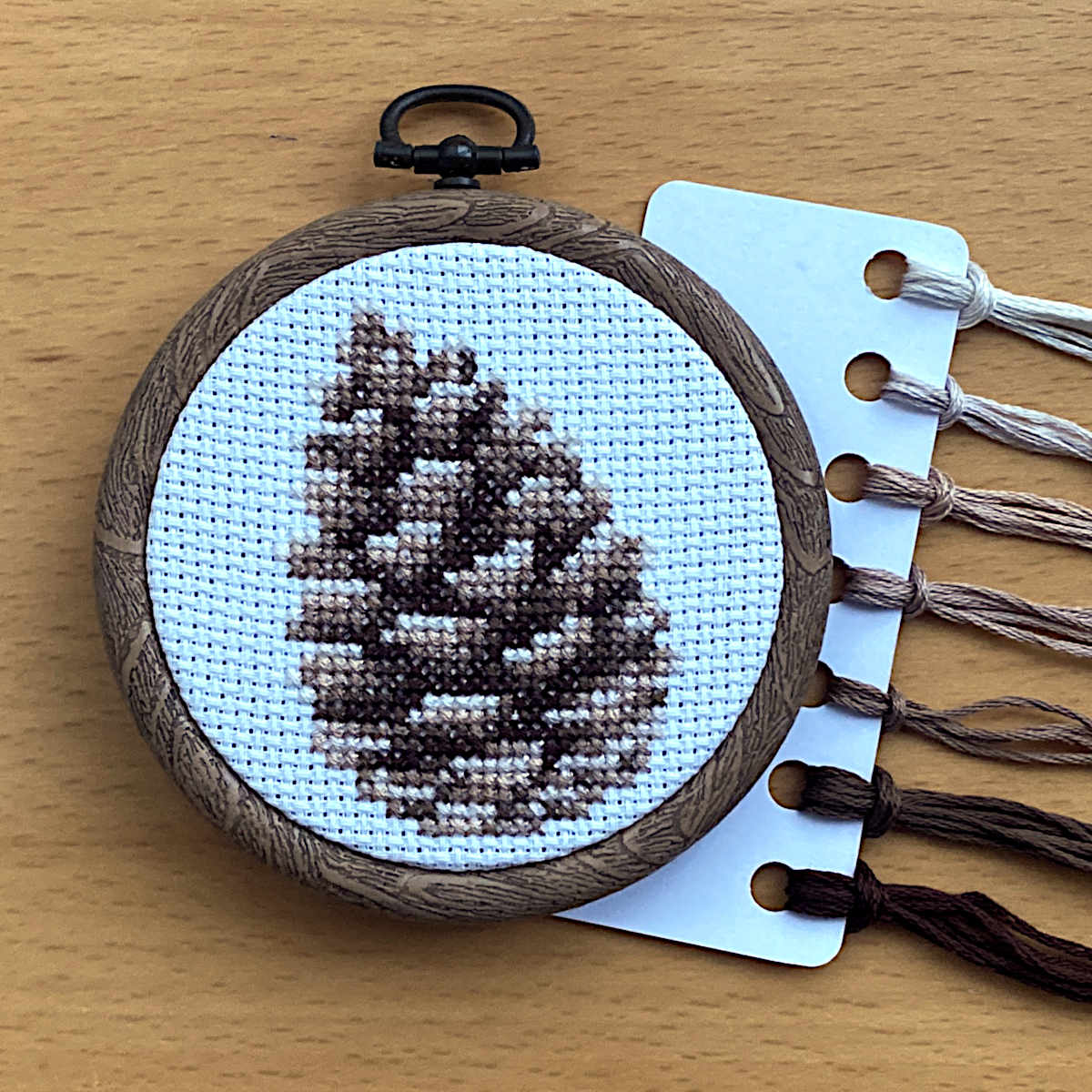 A completed pinecone cross stitch on white fabric, displayed in a round embroidery hoop with DMC threads