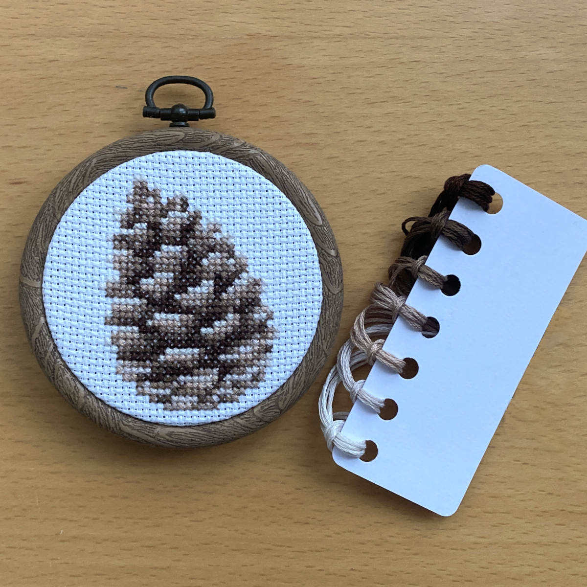 A completed pinecone cross stitch on white fabric, displayed in a round embroidery hoop with DMC threads