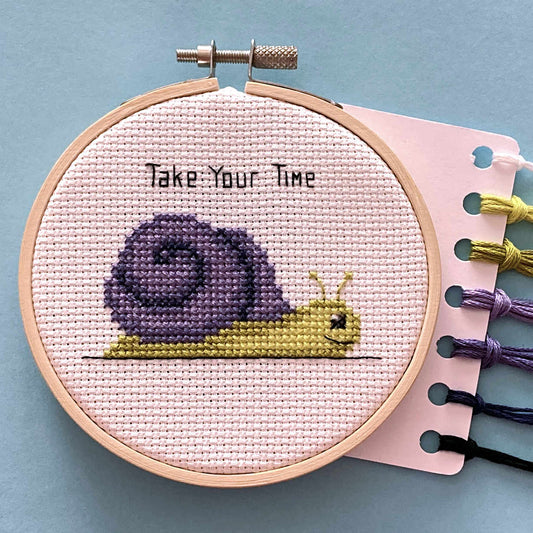 A completed cross-stitch design in a wooden hoop, featuring a purple and yellow snail with the text 'Take Your Time' stitched above it. A card with thread attached is visible on the right against a light blue background.