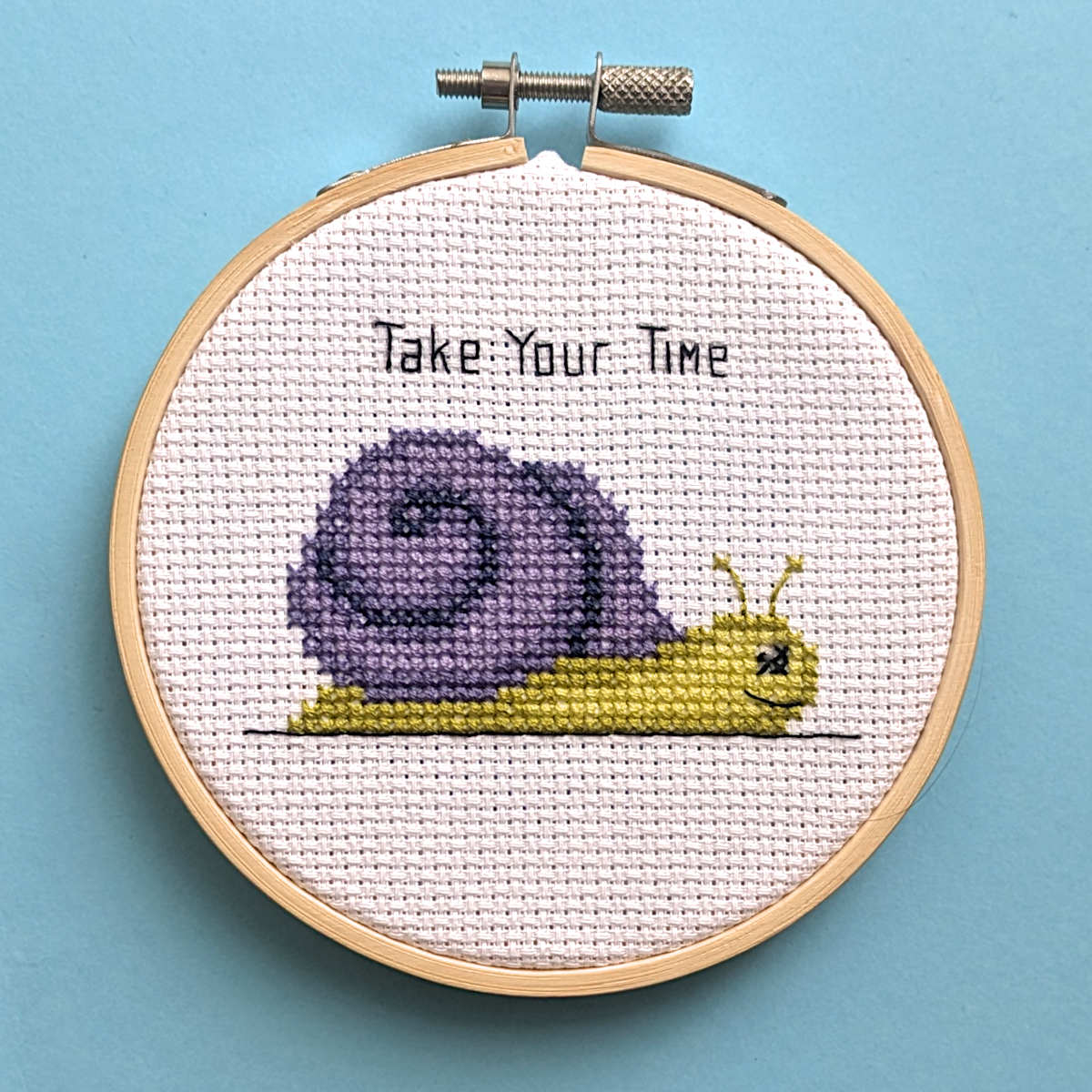 A completed cross stitch of a yellow-green snail with a purple shell with the backstitched caption 'Take Your Time' above it