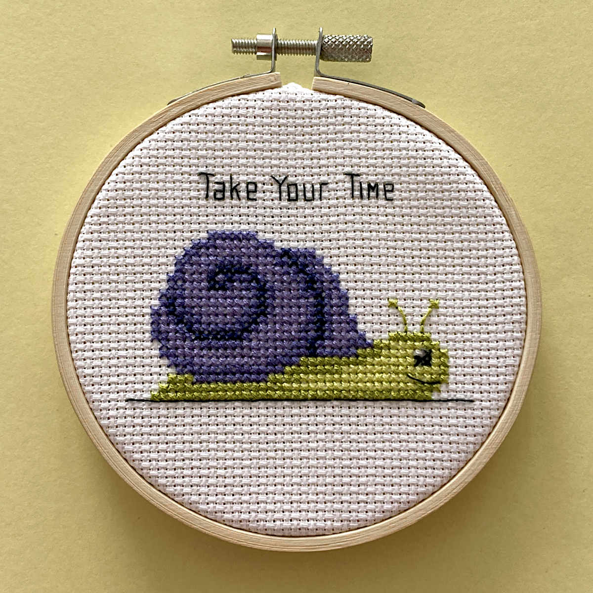 A circular embroidery hoop holding a white cross-stitch fabric with a purple and yellow snail and the words 'Take Your Time' stitched above it, set against a pale yellow background.