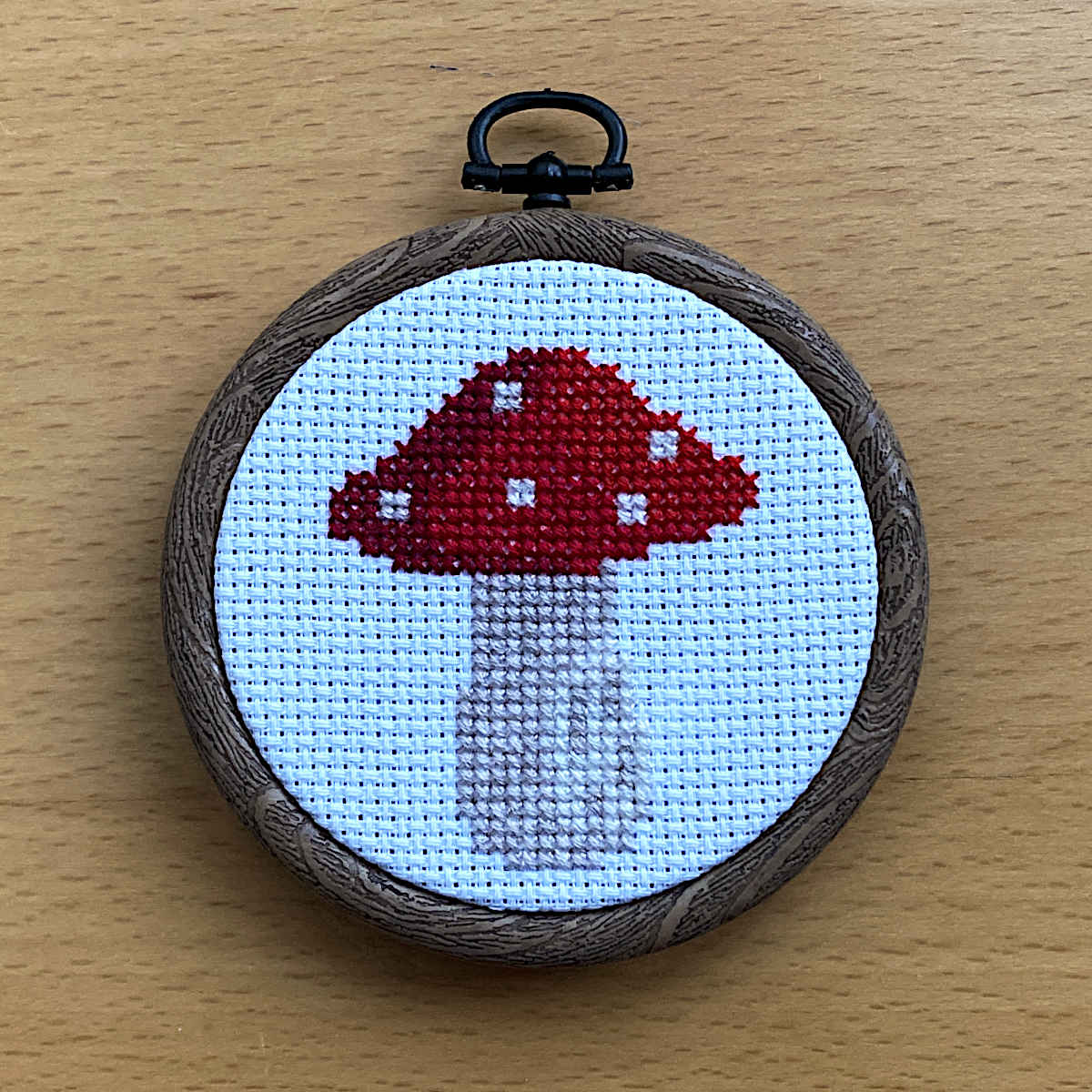 A circular cross-stitch embroidery hoop on a light wood surface, featuring a completed cross stitch of a mushroom design with a red cap, white spots, and a pale gray stem on a white fabric background.