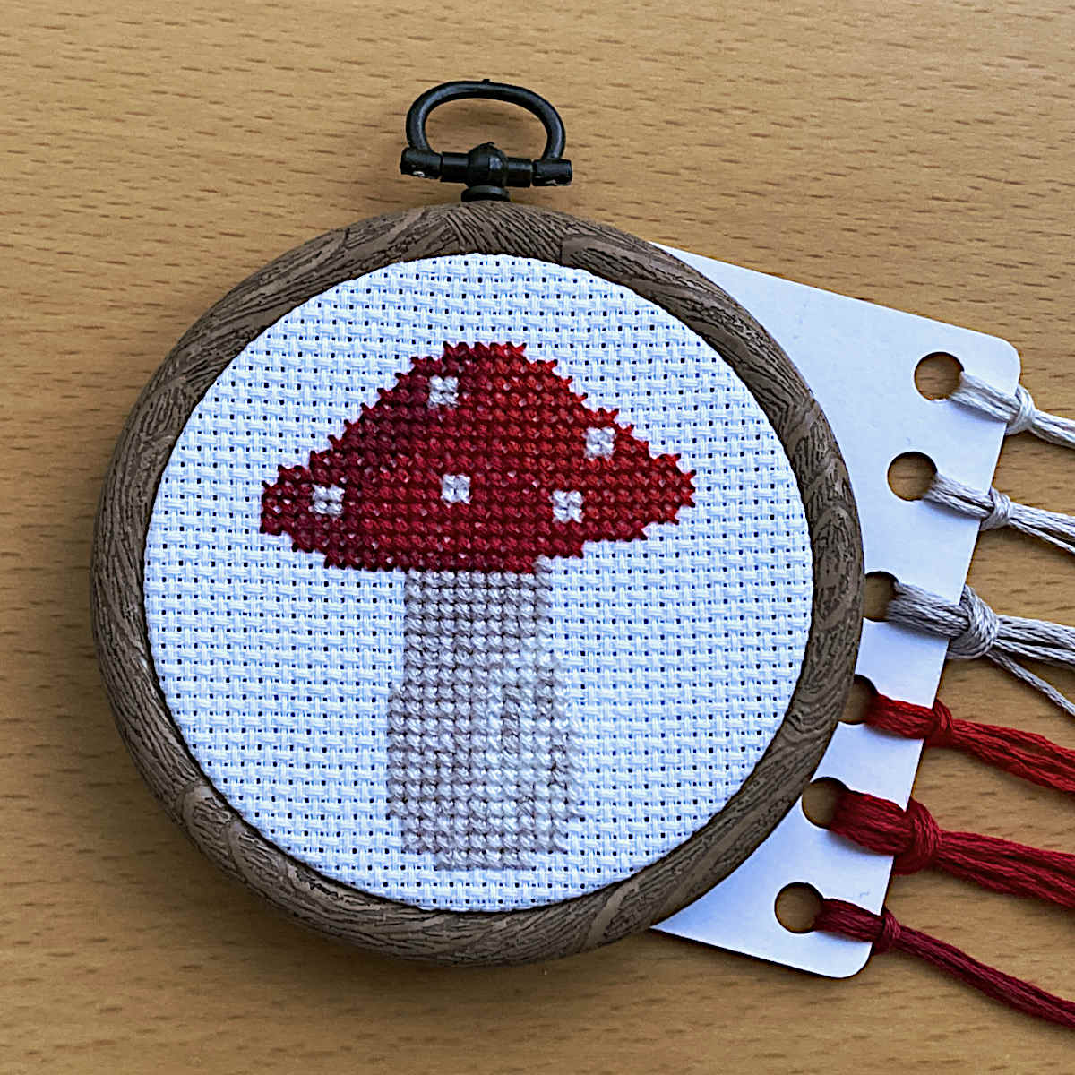 A small cross-stitch embroidery hoop featuring a red and white mushroom design on white aida fabric, displayed next to a card with colourful DMC threads.