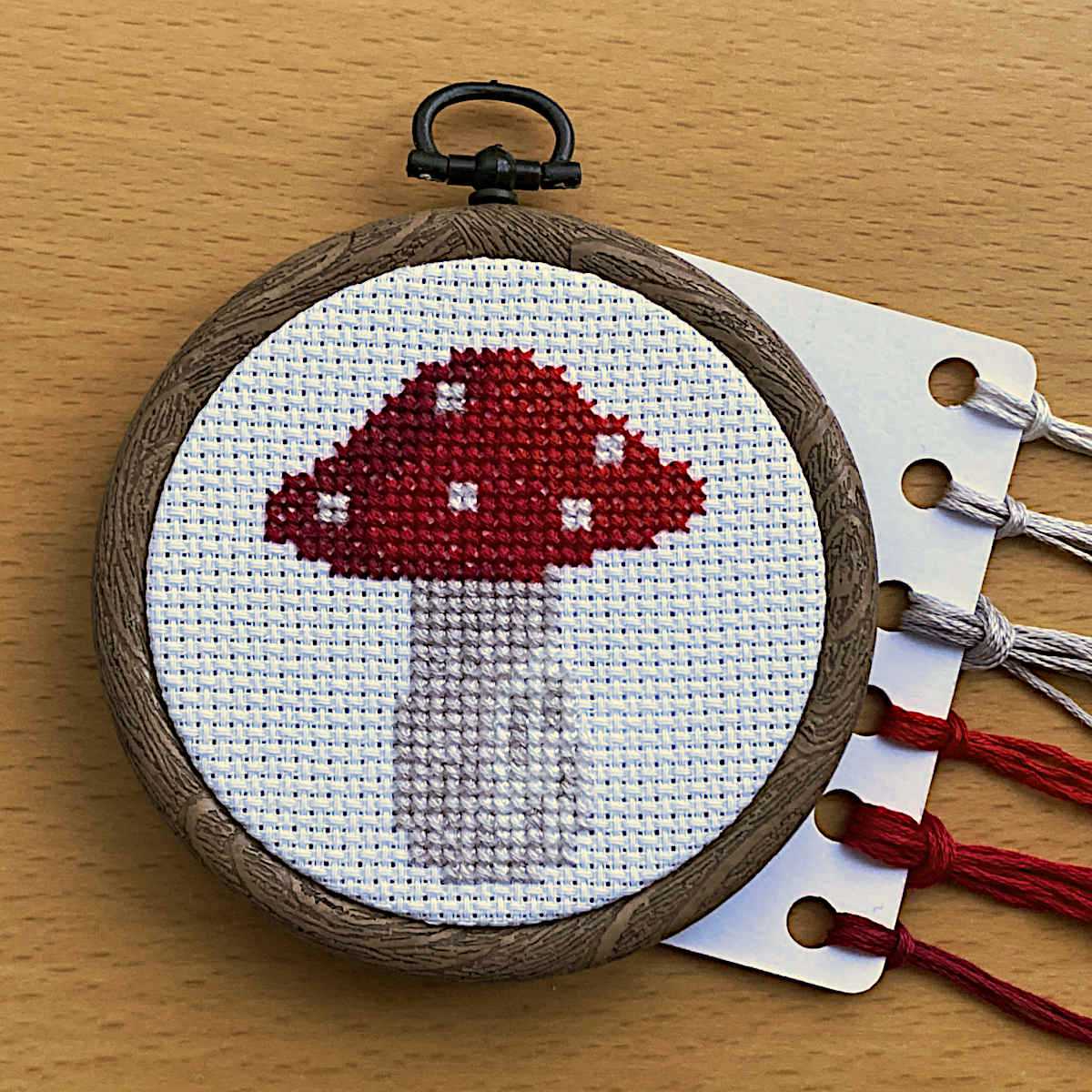 Cross stitch red-capped mushroom design with white spots, displayed in a round flexi embroidery hoop with DMC threads