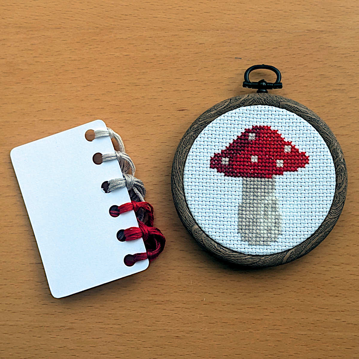 Cross stitch of a red capped toadstool with white spots, displayed in an embroidery hoop with DMC threads