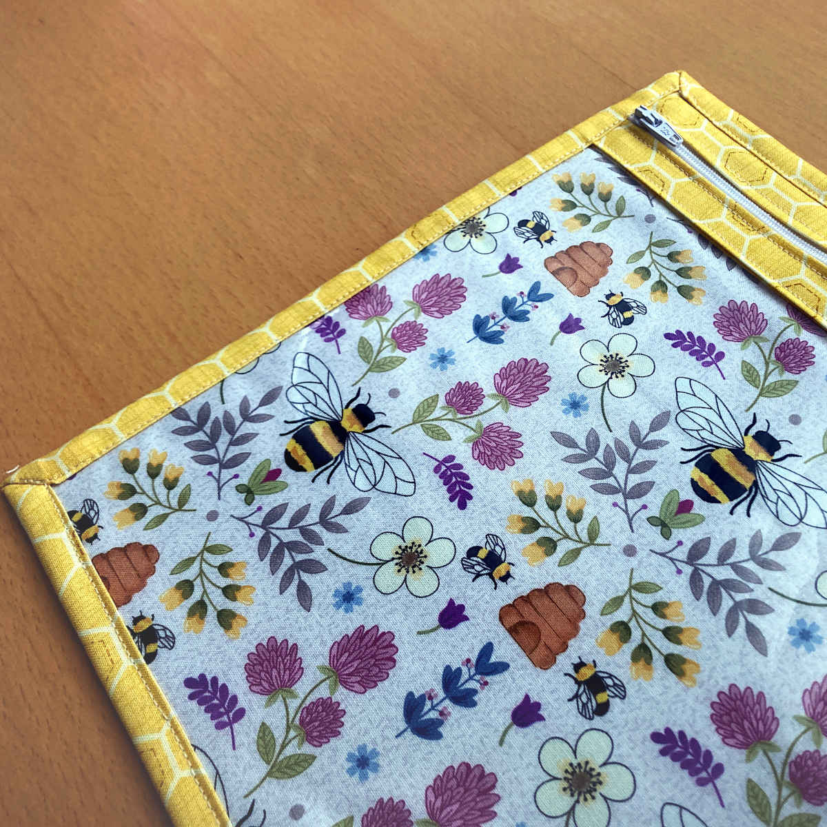 A close-up of a fabric zippered pouch with a yellow honeycomb outer fabric and zip and clear vinyl front and bee and flower print lining