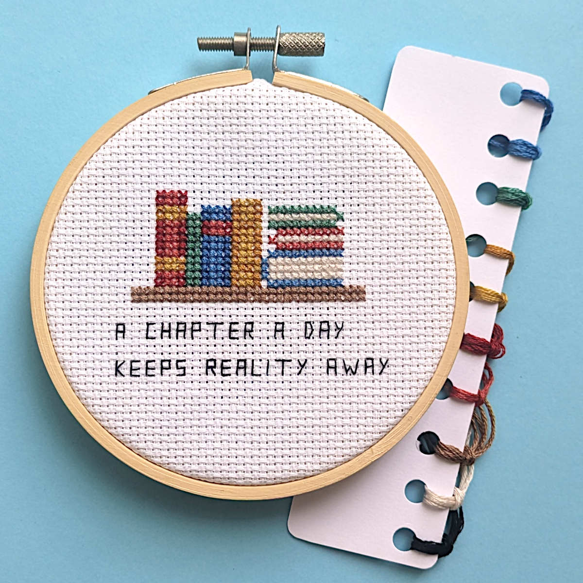 A completed cross stitch featuring books on a shelf and some stacked books with the caption 'A chapter a day keeps reality away' in backstitch. Displayed in a 4in embroidery hoop with DMC thread card next to it.