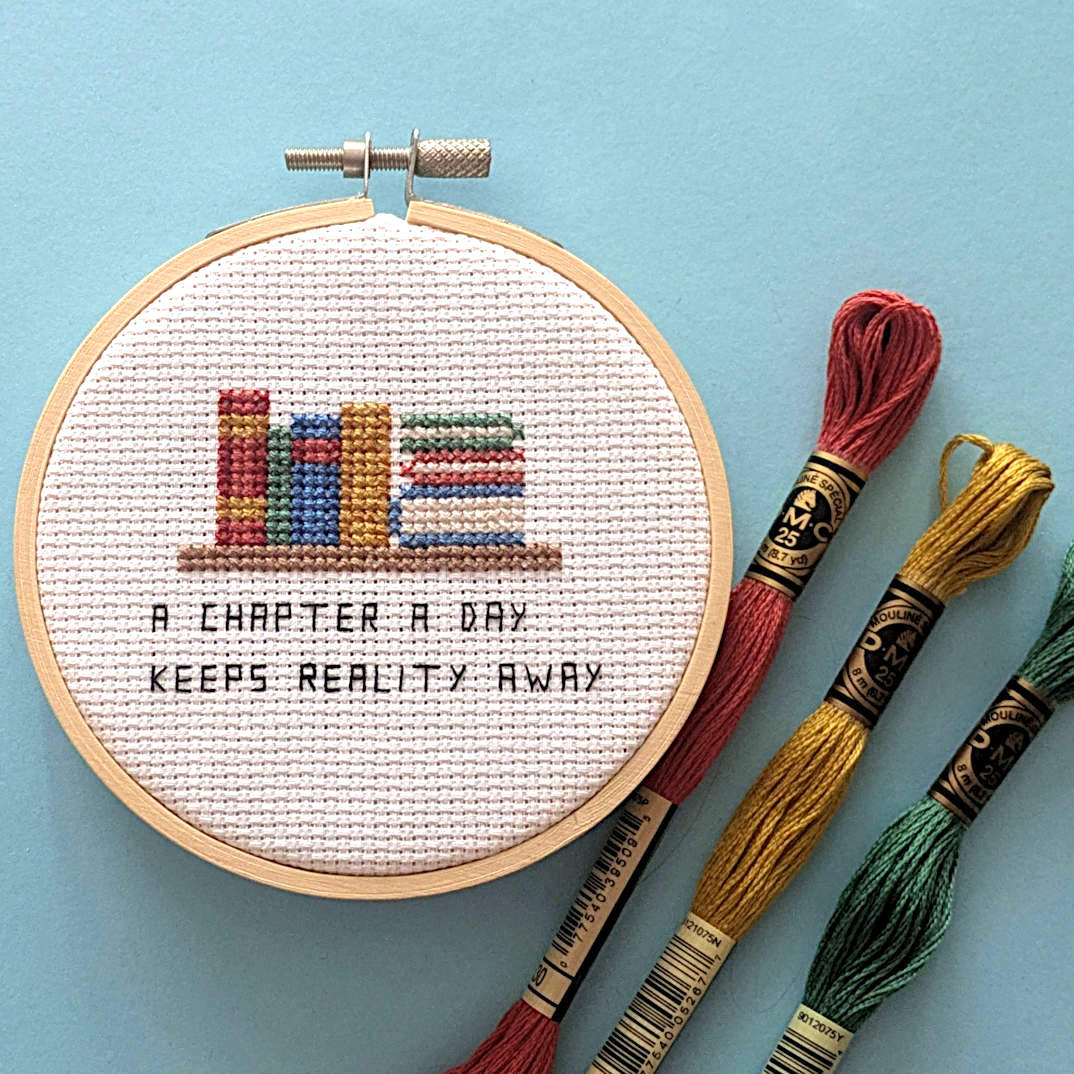 A completed cross-stitch in a wooden hoop showing colorful books on a shelf and the text 'A CHAPTER A DAY KEEPS REALITY AWAY', displayed on a light blue background with three embroidery floss skeins beside it.