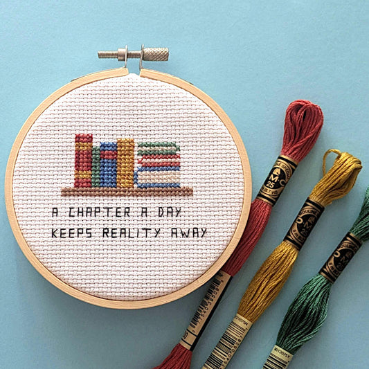 A completed cross-stitch in a wooden hoop showing colorful books on a shelf and the text 'A CHAPTER A DAY KEEPS REALITY AWAY', displayed on a light blue background with three embroidery floss skeins beside it.