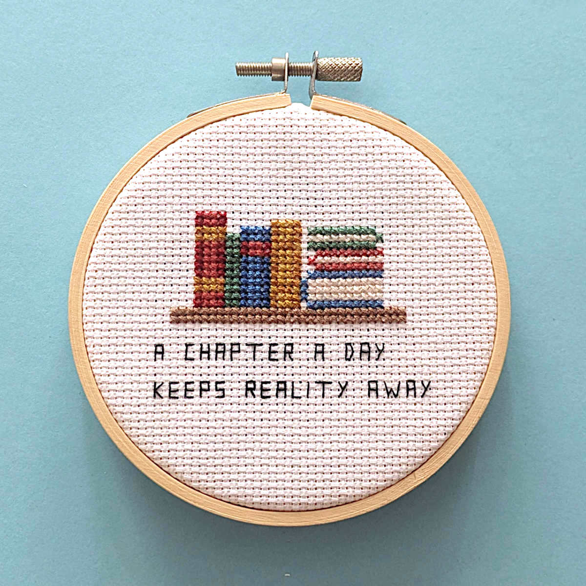 A finished cross-stitch embroidery piece in a wooden hoop, featuring colorful books on a shelf and the text 'A CHAPTER A DAY KEEPS REALITY AWAY', set against a solid light blue background.