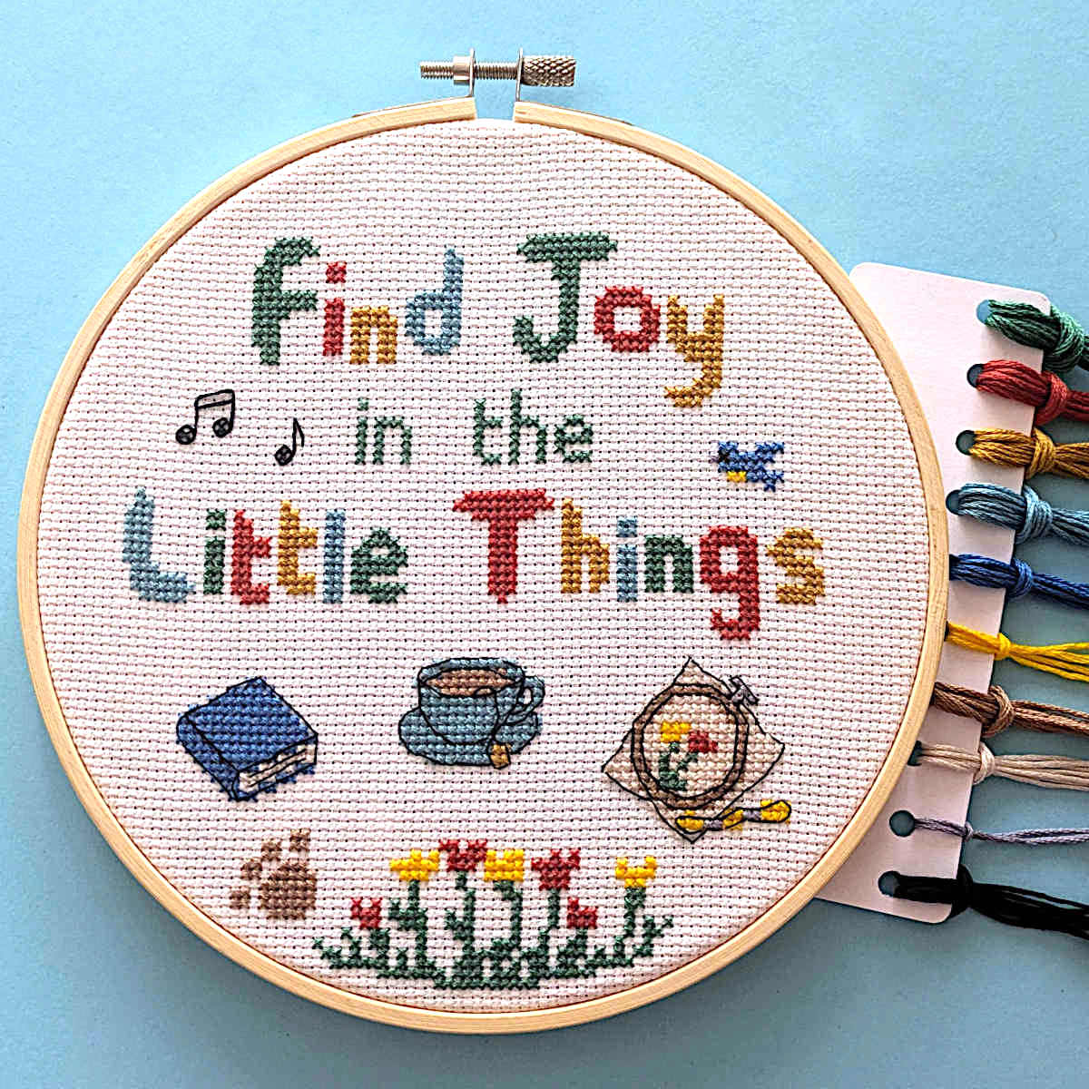 A completed cross-stitch embroidery in a wooden hoop on a light blue background, with a card of colorful DMC embroidery floss threads to the right. The text reads 'Find Joy in the Little Things' with small stitched icons including music notes, a book, a cup of tea, a paw print, an embroidery, a bird and flowers.