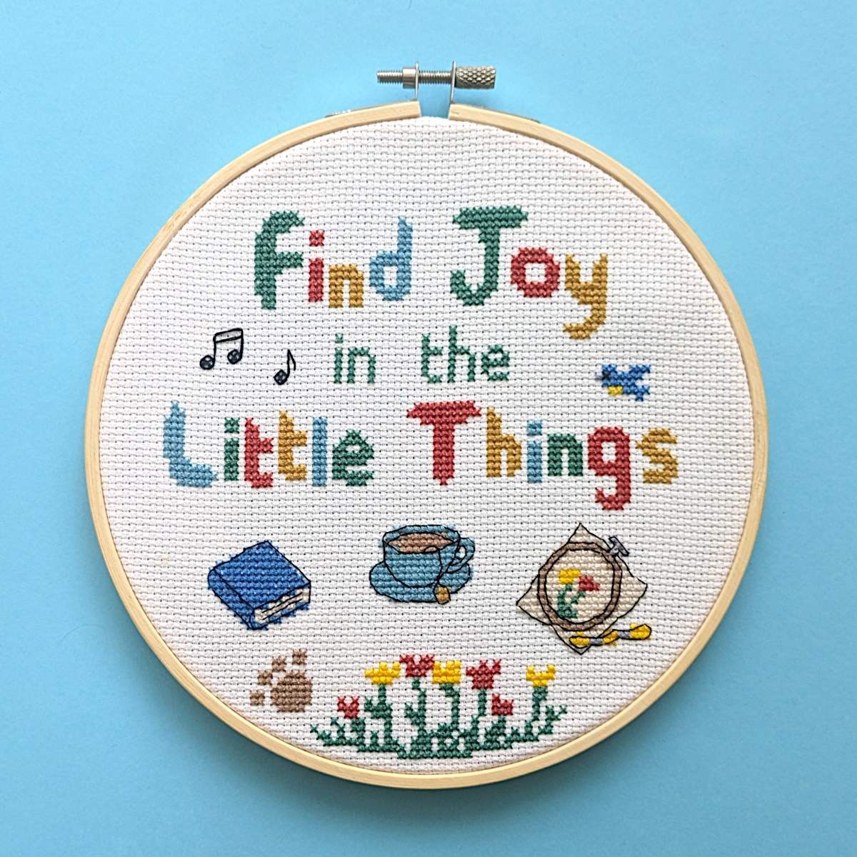 A round wooden embroidery hoop holding a cross-stitch design that reads 'Find Joy in the Little Things' with colorful stitched letters and small motifs including music notes, a book, a bird, a cup of tea, an embroidery hoop, flowers, and a paw print, set against a solid light blue background.