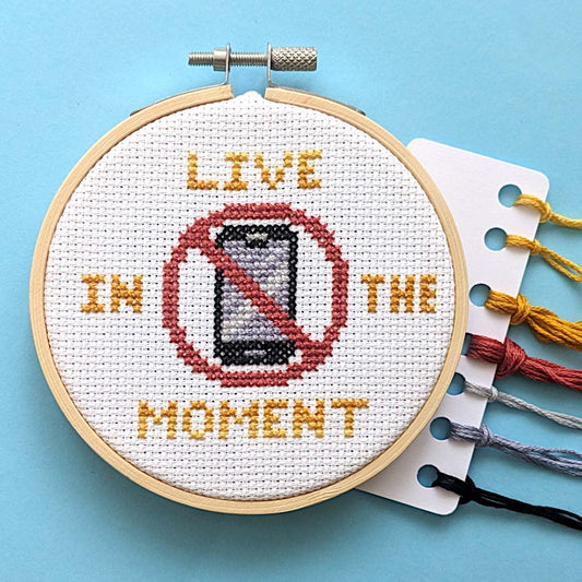 A completed cross-stitch embroidery in a wooden hoop on a light blue background. The design reads 'LIVE IN THE MOMENT' with a red prohibition symbol over a smartphone.