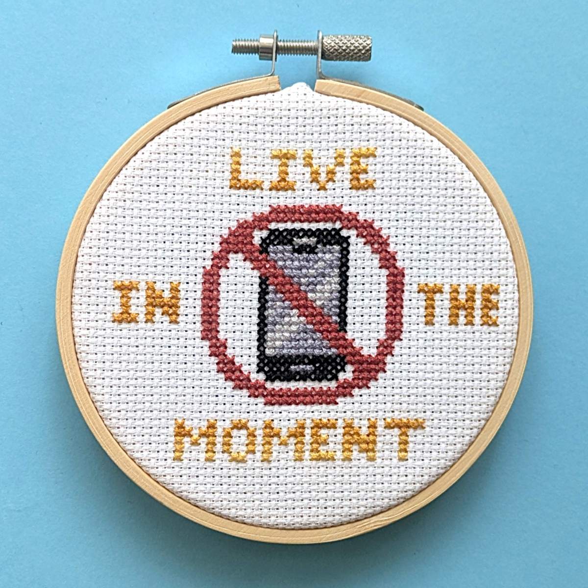 A circular embroidery hoop on a light blue background, featuring completed cross stitch design with the words 'LIVE IN THE MOMENT' and a red prohibition symbol over a smartphone graphic.