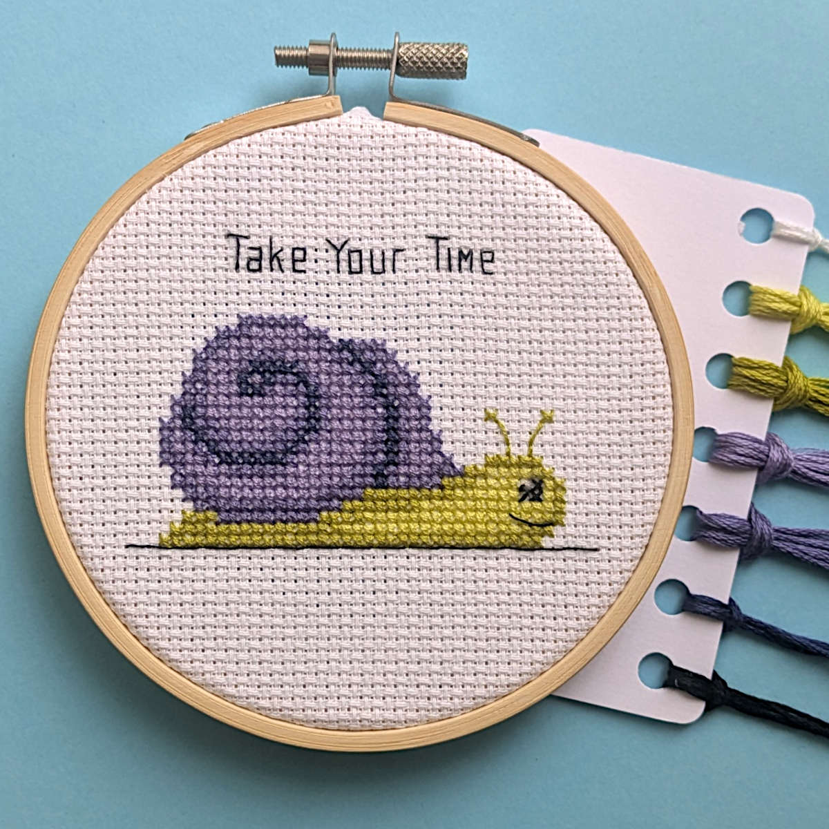 A completed cross stitch of a yellow-green snail with a purple shell with the backstitched caption 'Take Your Time' above it. Displayed in a 4in embroidery hoop with DMC threads to the side.